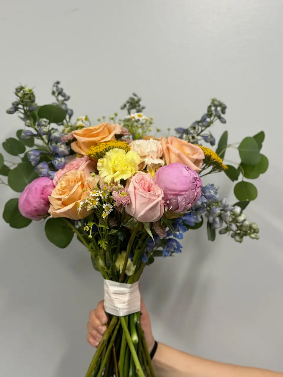 A hand holding up a bright bouquet of pink, peach and yellow blooms with greenery